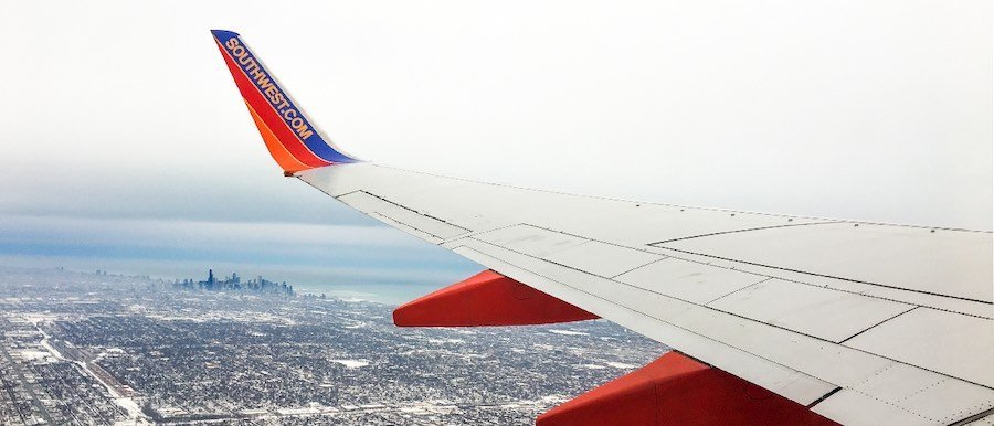 Photograph from a Soutwest aircraft looking out the left side of the plane over the wing. Below is a city in the horizon with the suburbs surrounding it covered in snow.